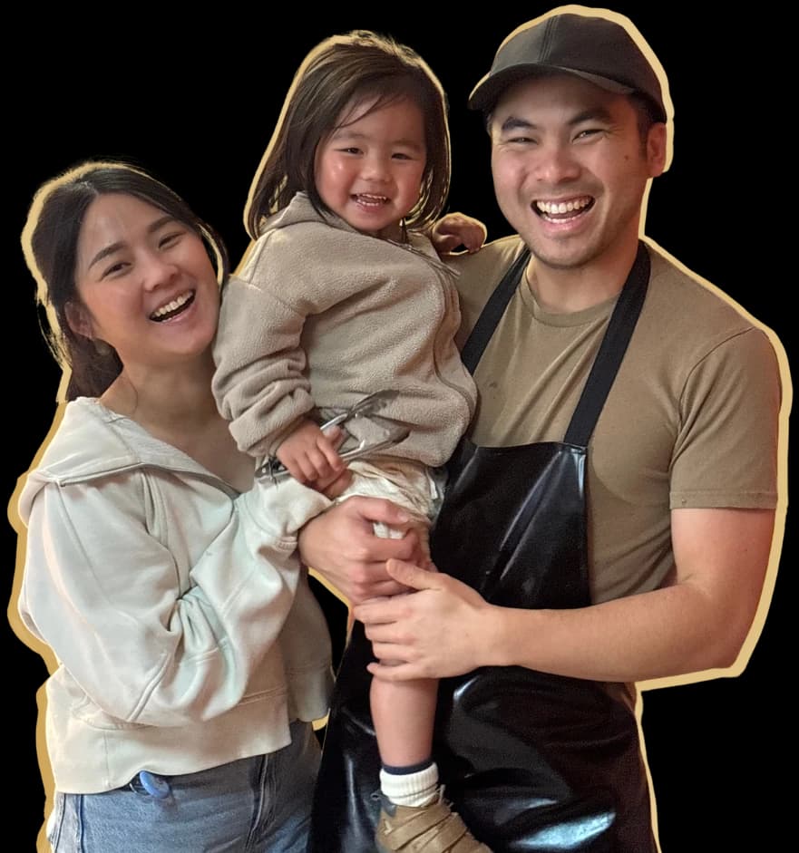 The family who own Kaiapoi Roast: the owner in his cap and apron holding a young child, with a smiling woman beside them, all laughing